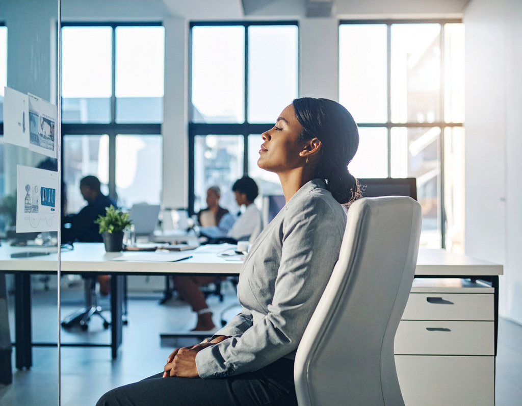 Image shows a woman sitting at her desk at work, practising mindfulness and looking peaceful